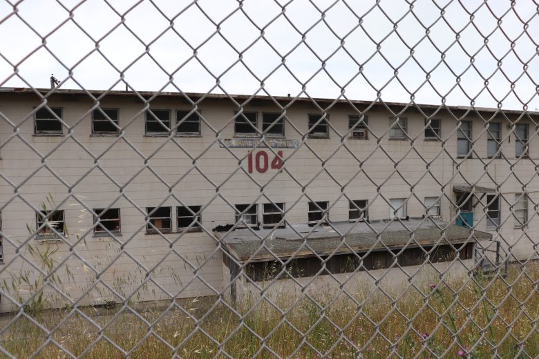 An abandoned, two-story building with broken windows and a chain-link fence in front; the number "104" is displayed above a doorway.