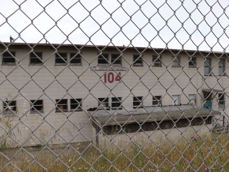 An abandoned, two-story building with broken windows and a chain-link fence in front; the number "104" is displayed above a doorway.