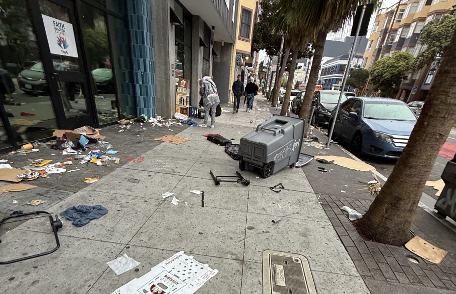 Litter and debris scattered across a city sidewalk with overturned trash bins, a few people walking, and parked cars along the street.