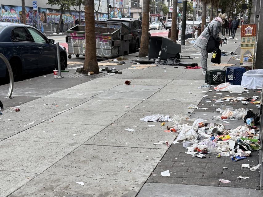 Sidewalk littered with trash, food containers, and paper debris; a person with bags stands near crates, and a dumpster is in the background beside parked cars and palm trees.