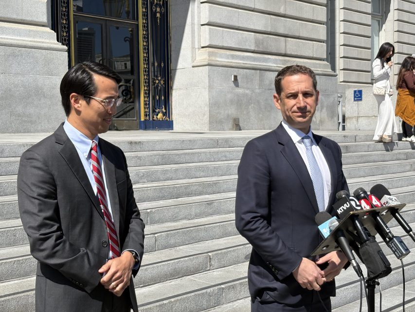Two men in suits stand near a podium with multiple microphones in front of a building with stone steps. One man is speaking, and the other is smiling while looking on.