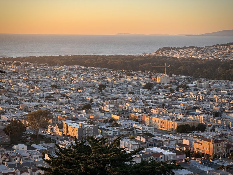 Aerial view of a city at sunset, with densely packed buildings and a forested area leading to the ocean in the background.