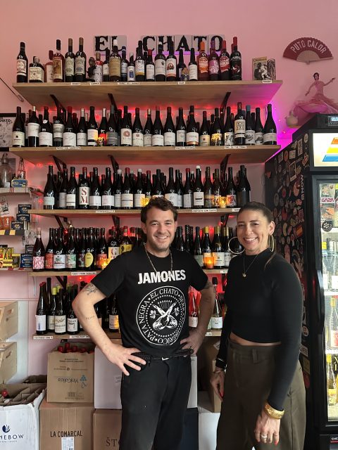 Two people stand in front of shelves brimming with bottles of wine. The person on the left dons a "Jamones" shirt, while the one on the right wears black. They both seem captivated by an El Chato wine selection, sharing a quiet moment in this SF cellar sanctuary.