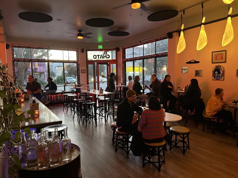 Patrons enjoy El Chato wine as they sit at tables and the bar in a cozy, dimly lit café with large windows. Warm lighting and wooden floors create an inviting atmosphere, echoing the vibrant yet laid-back spirit of SF.