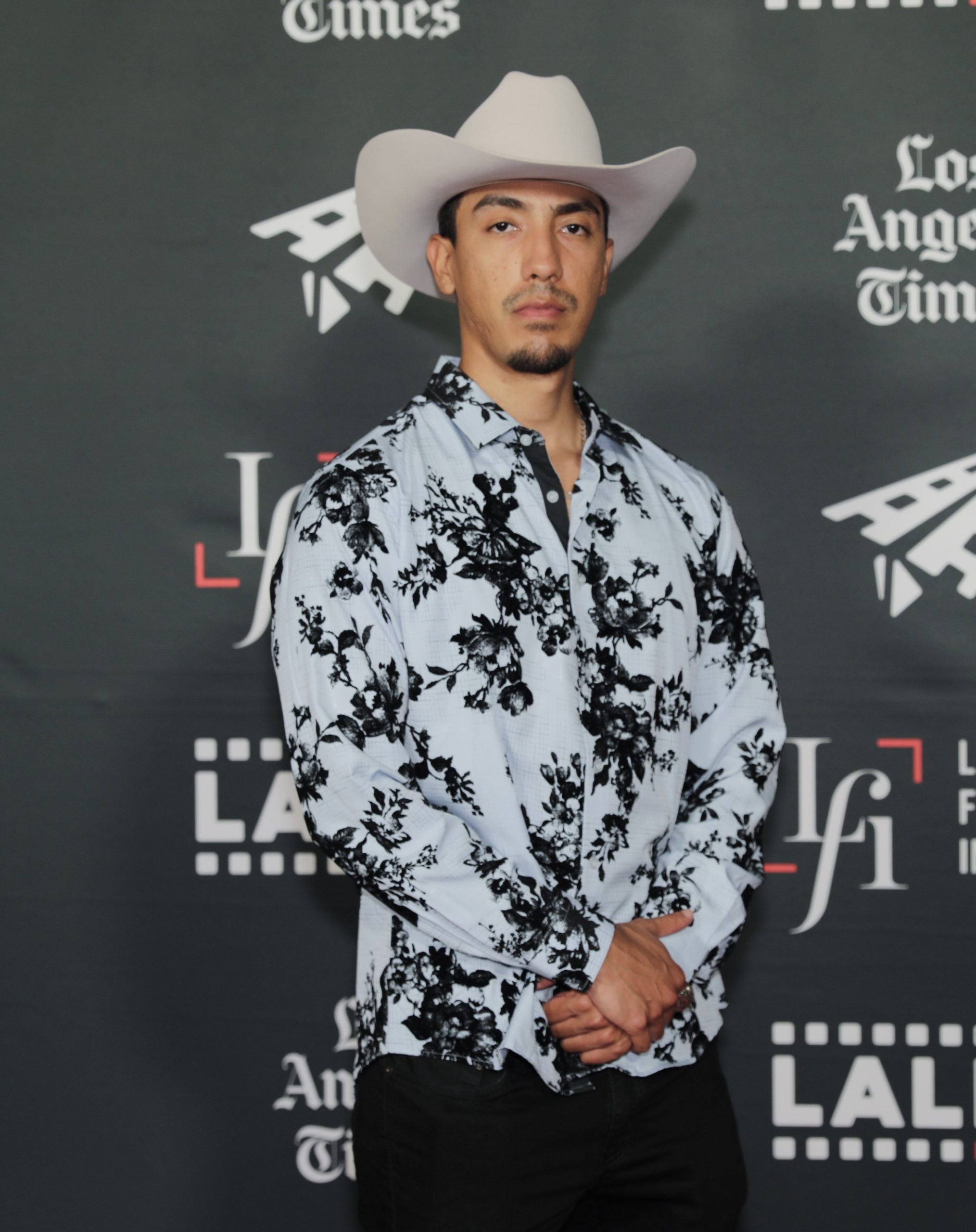 A man in a white cowboy hat and floral shirt stands confidently in front of a backdrop adorned with Los Angeles Times logos, embodying the playful yet bold spirit of Roxie.