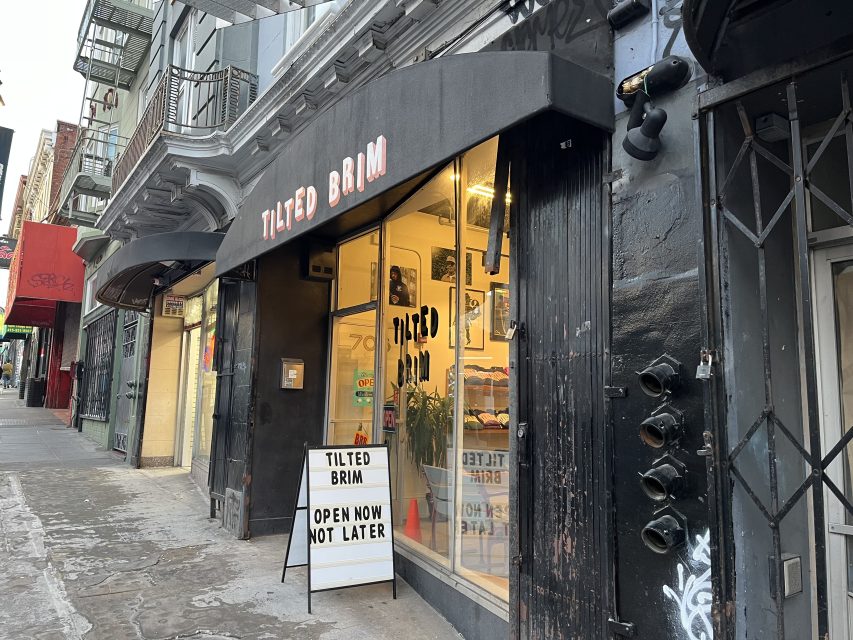 Street view of Tilted Brim storefront with a black awning and a sign that reads, “OPEN NOW NOT LATER.” The shop window is lit and displays merchandise inside.