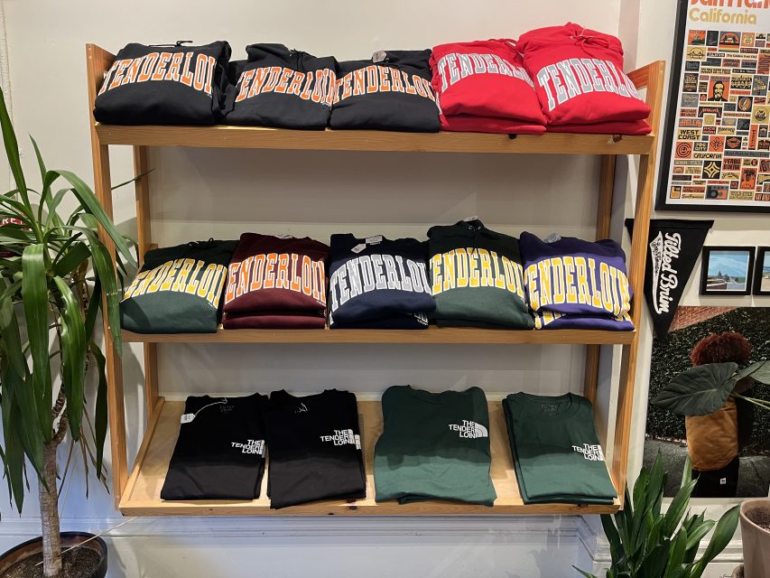 A wooden shelf displays folded “TENDERLOIN” sweatshirts in various colors on the top two shelves and green and black t-shirts with small text designs on the bottom shelf.