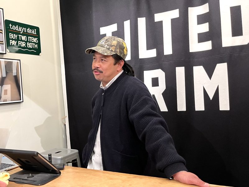 A man in a dark jacket and camouflage cap stands behind a counter near a tablet, with a black wall that reads Tilted Brim.