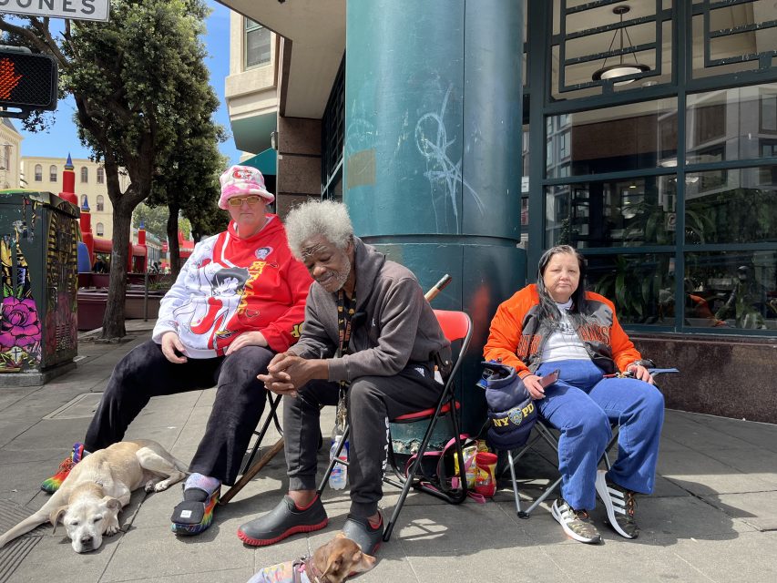 Three people seated on folding chairs by a street corner with two dogs lying on the ground. A traffic signal and street signs are visible in the background.