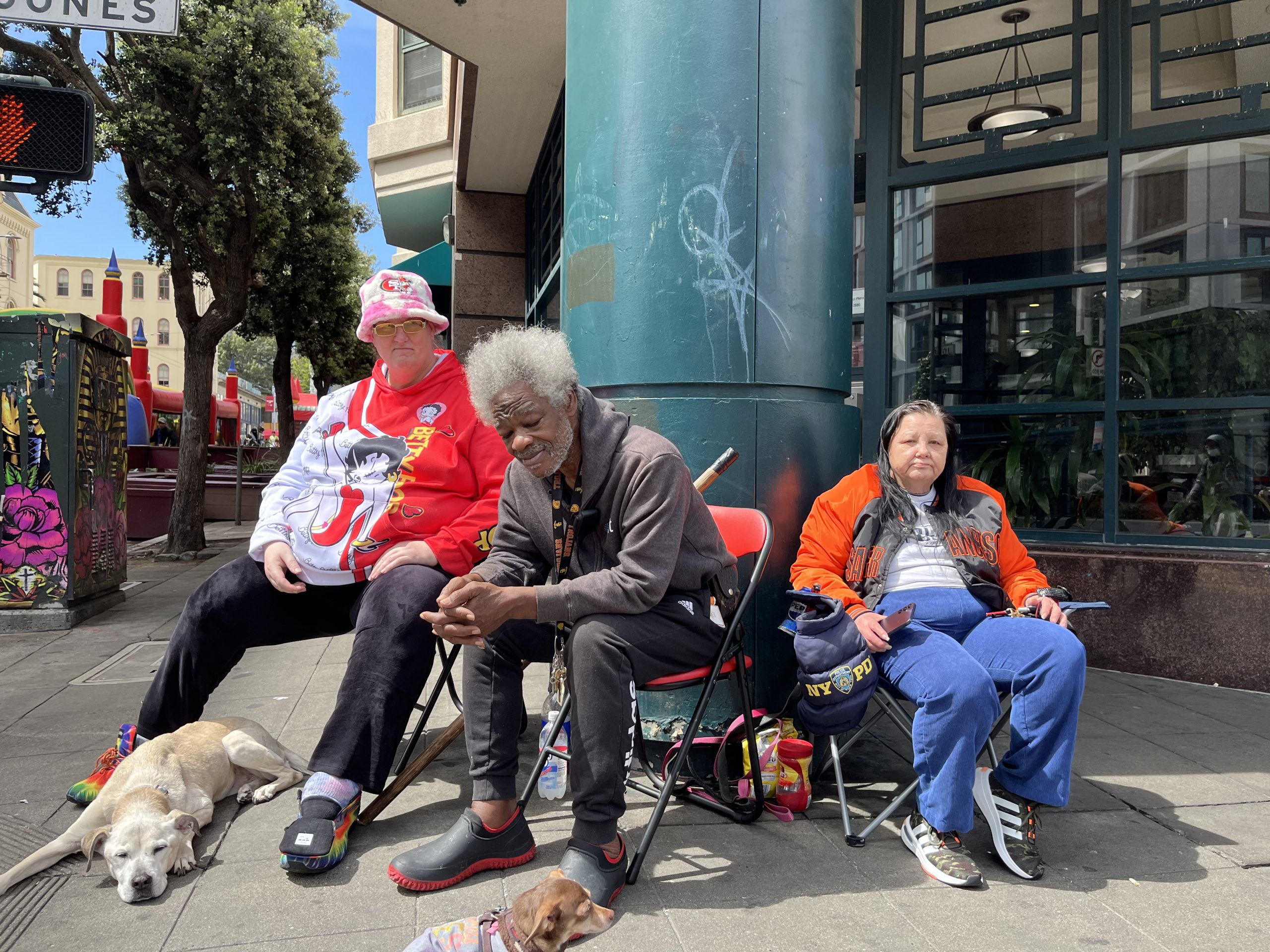 Three people seated on folding chairs by a street corner with two dogs lying on the ground. A traffic signal and street signs are visible in the background.