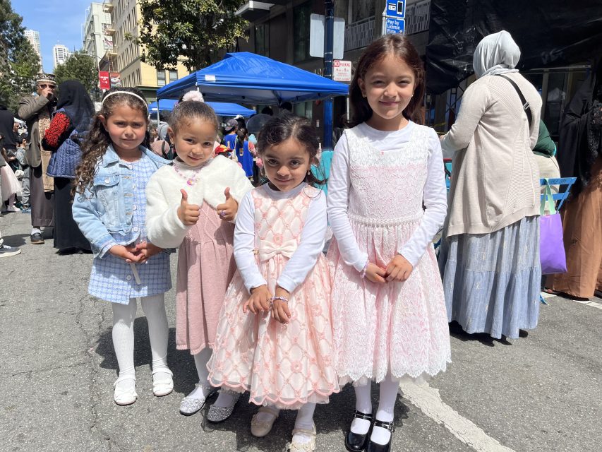 Four young girls in dresses pose together outdoors at a street event, with a blue canopy and people in the background.
