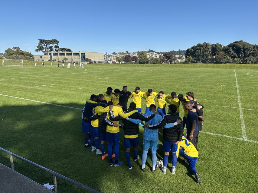 A soccer team in yellow jerseys stands in a huddle on a grassy field, while another group practices in the background under a clear blue sky.