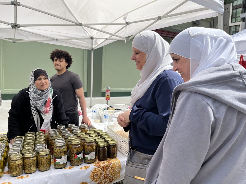 People at a market stall with jars of pickled items. Two women in hijabs are engaged in conversation with the vendor, and a man stands nearby.