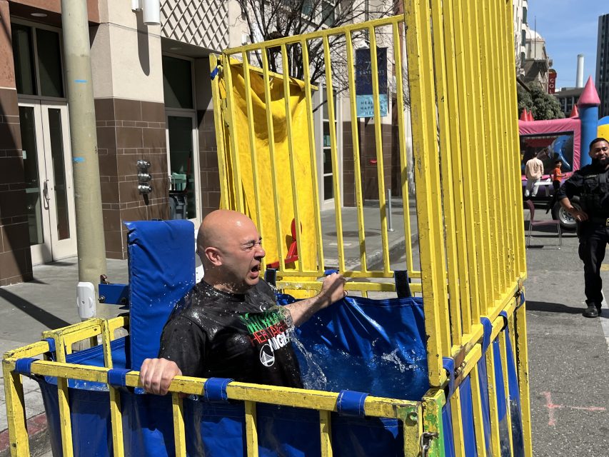 A person is sitting in a dunk tank after being dropped into the water, with water splashing around. A person in the background is looking on.