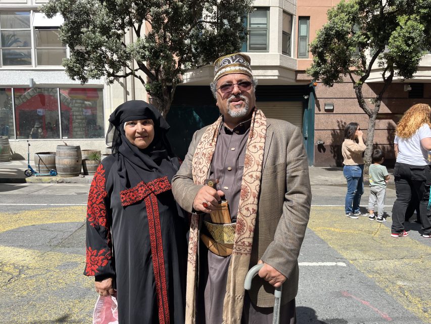 A man and woman in traditional attire stand on a city street during the day. The man holds a cane. Trees and buildings are visible in the background.