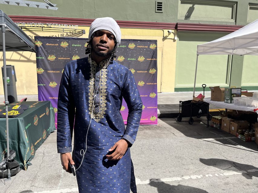 Man wearing a blue, gold-trimmed traditional outfit and a white head covering stands outdoors on a sunny street. A branded backdrop and canopies are in the background.