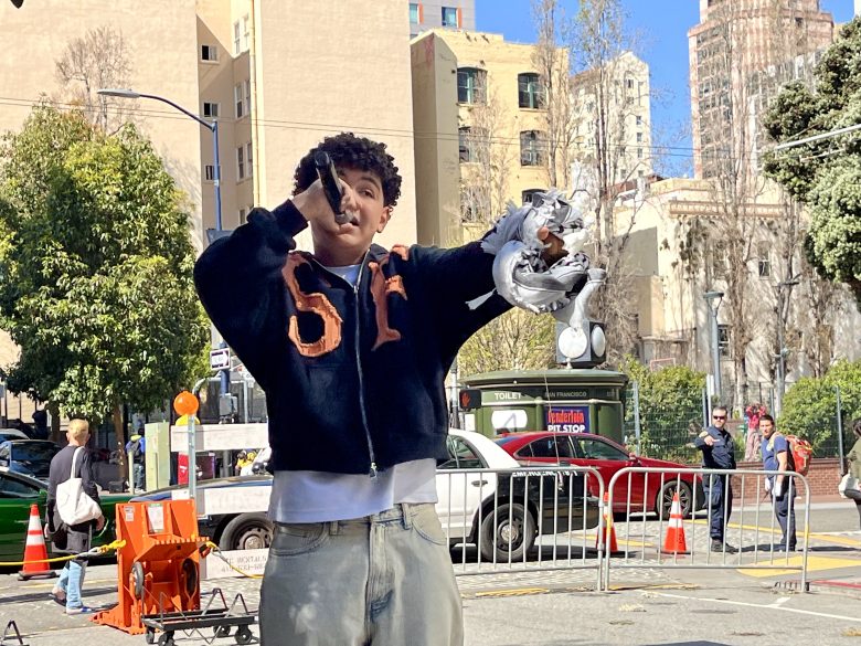 Person in a dark jacket and light pants sings into a microphone on a city street. There are buildings, trees, and spectators in the background.
