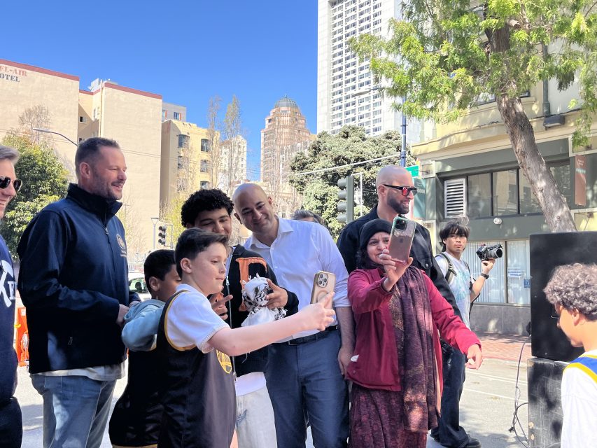 A group of people, including three children and several adults, take a selfie on a city street. Trees and buildings are visible in the background.