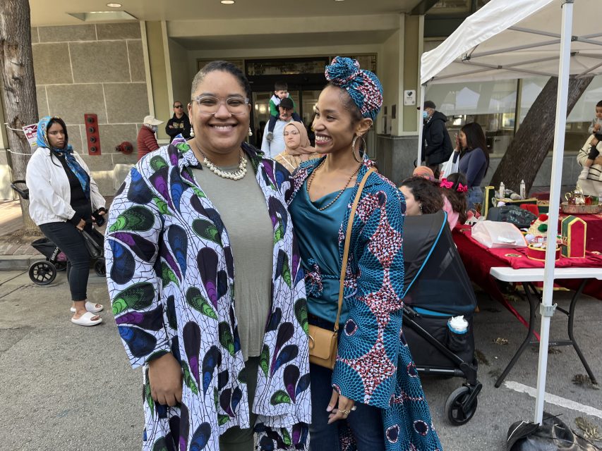 Two people smiling and standing outdoors at an event. Both wear colorful, patterned clothing. There's a tent with items and people in the background.