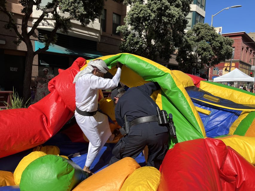 A person in a karate gi and a police officer help deflate a colorful bounce house on a city street.