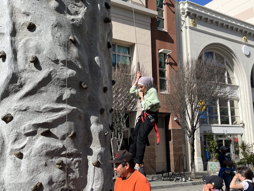 Child wearing a helmet and harness climbs a rock wall outdoors with an adult supporting. Buildings in the background.