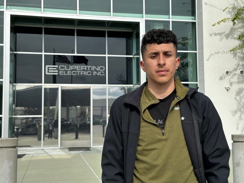 A person stands in front of the Cupertino Electric Inc. building, which has large glass windows and a visible company logo above the entrance.