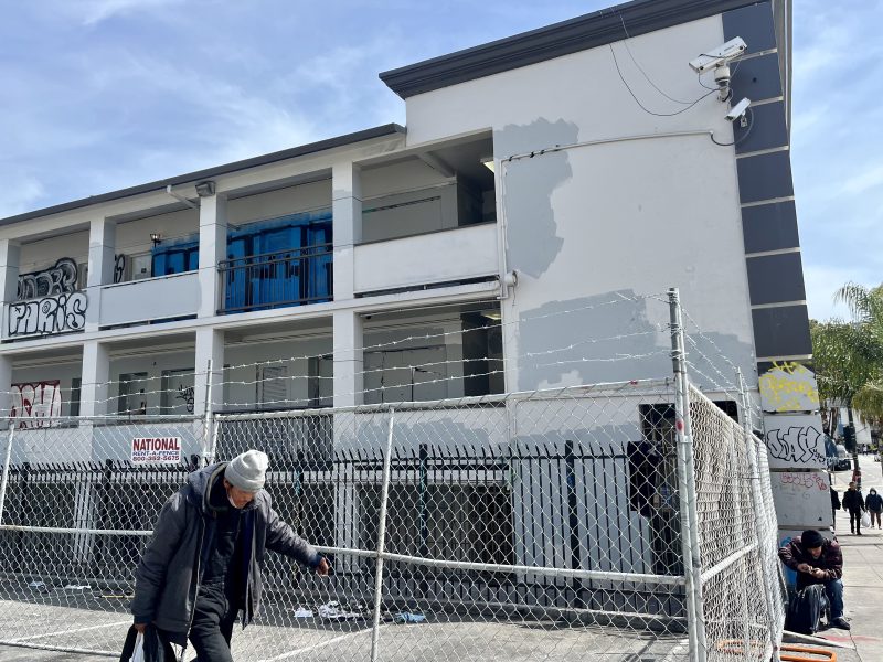 A person walks past a barbed wire fence in front of a two-story building with partially painted walls, graffiti, and security cameras.