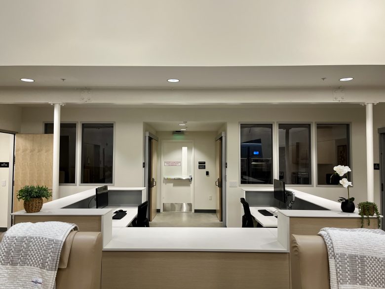 A medical office reception area with two workstations, computer monitors, chairs, a sink in the background, and potted plants on the counter.