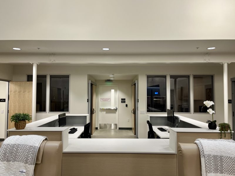 A medical office reception area with two workstations, computer monitors, chairs, a sink in the background, and potted plants on the counter.