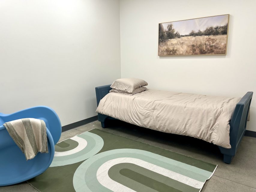 A simple bedroom with a single bed, beige bedding, a green patterned rug, a blue chair with a draped blanket, and a landscape painting on a white wall.