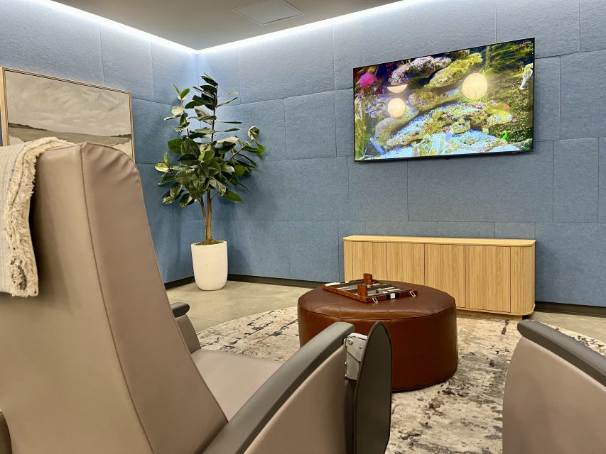 A modern lounge with two gray chairs, a round brown ottoman with a tray, a potted plant, a wall-mounted TV displaying nature, and light blue walls.