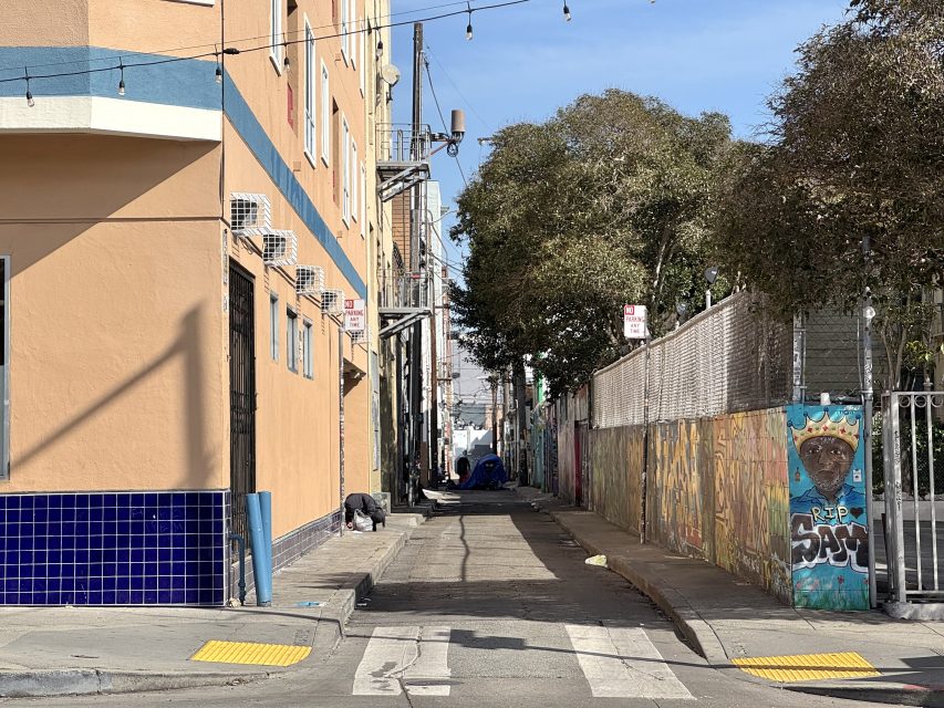 Narrow alley with graffiti, trees, and overhead string lights. A person leans against a peach-colored building on the left. Blue-tiled section at the building’s bottom.
