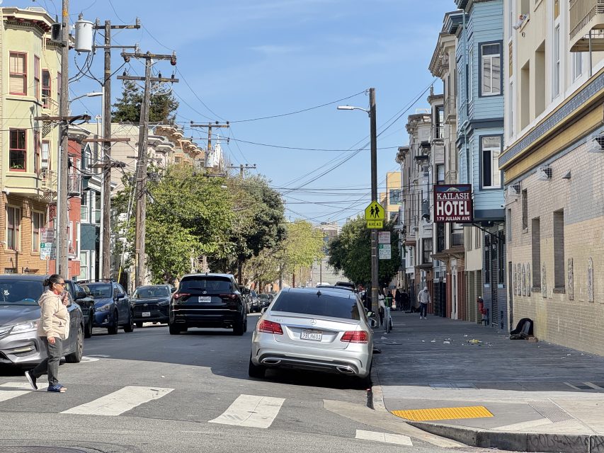 A street scene with parked cars, a person crossing the street, and a hotel sign on a sunny day.