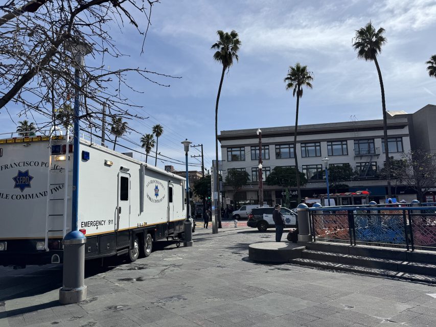 A police mobile command unit is parked in a plaza with palm trees and a building in the background under a partly cloudy sky.