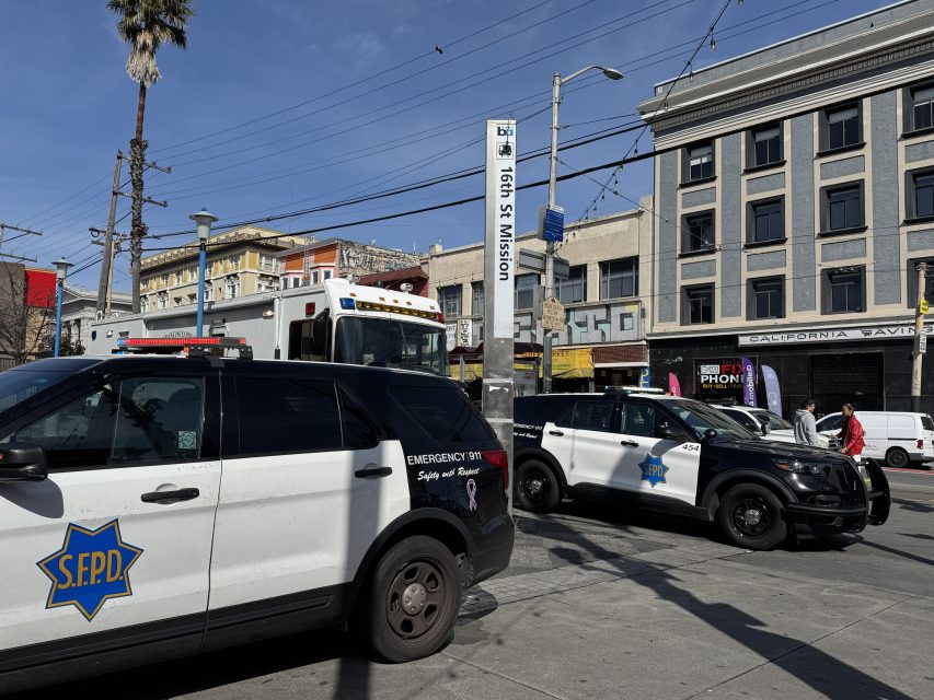 Two police SUVs parked near the 16th St Mission station sign in a city setting with buildings and a fire truck in the background.