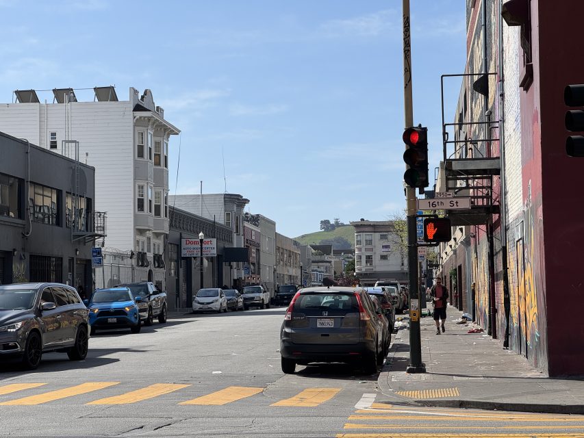 Street intersection on a sunny day with cars parked along the sides, a stoplight showing red, and buildings in the background. Crosswalk and pedestrian visible in the scene.