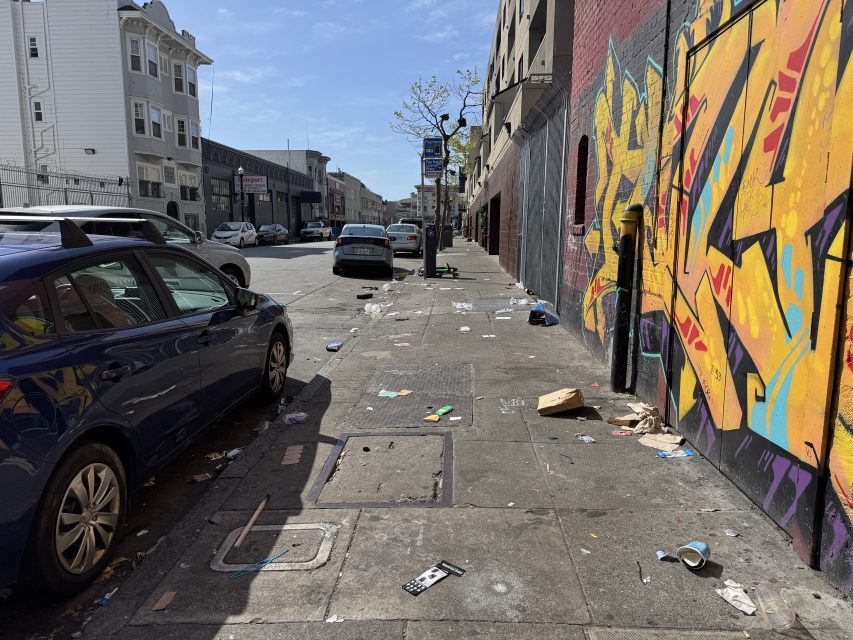Urban street with parked cars, colorful graffiti on a wall, and litter scattered on the sidewalk.