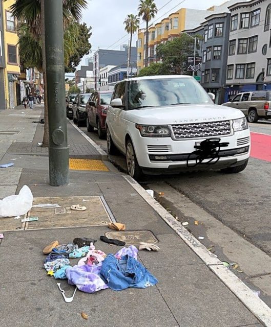 Clothes, shoes, and personal items are scattered on a city sidewalk next to a white SUV parked along the street.