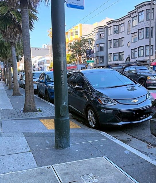 A black Chevrolet Bolt is parked at the curb on a city street lined with palm trees and multi-story buildings.