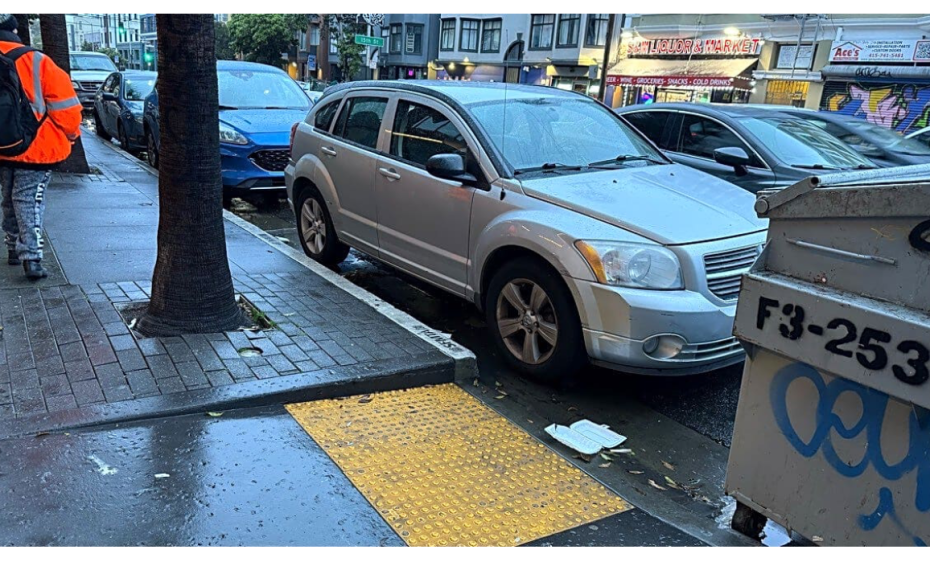 A silver car is parked blocking a sidewalk curb ramp on a wet city street beside a dumpster and a tree.