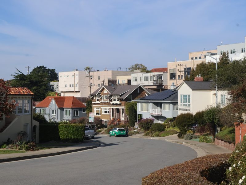 A residential street with houses on both sides, a green and white vehicle on the road, and a clear blue sky above.