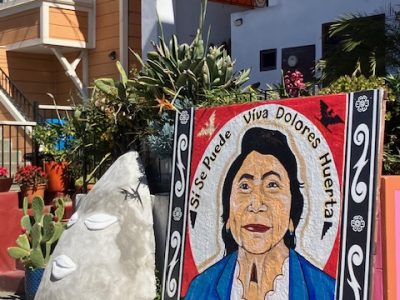 A mural of a woman with "Sí Se Puede Viva Dolores Huerta" inscribed, set in a garden with plants, cacti, and a white sculpture nearby.