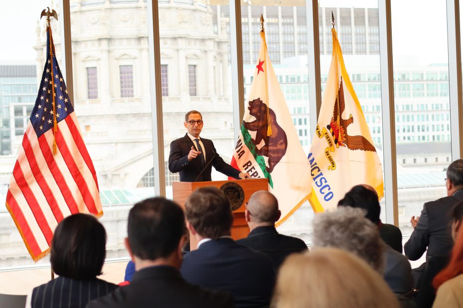 A man speaks at a podium in front of an audience, with the American, California, and San Francisco flags displayed behind him in a large room with tall windows.