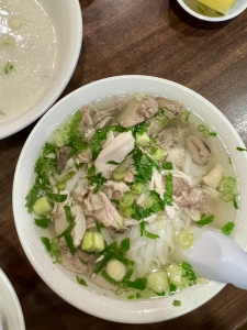 A bowl of Vietnamese pho with sliced green onions, cilantro, chicken pieces, and rice noodles in clear broth, on a wooden table.