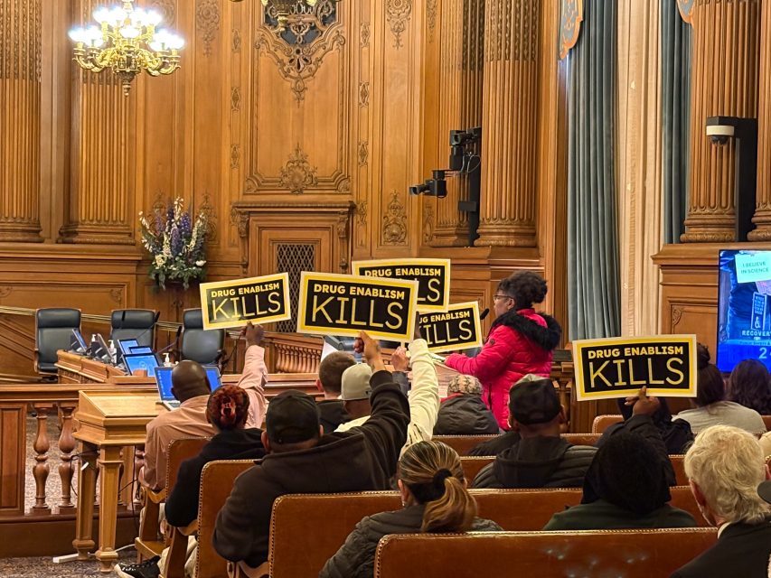 People seated in a wood-paneled meeting room hold up signs reading "DRUG ENABLISM KILLS" during what appears to be a public hearing or meeting.