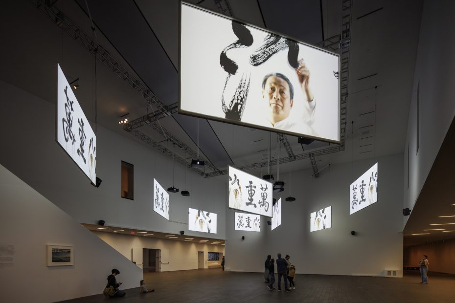 Large video screens display a person painting calligraphy and finished calligraphic works in a modern, spacious museum gallery with visitors observing the installation.