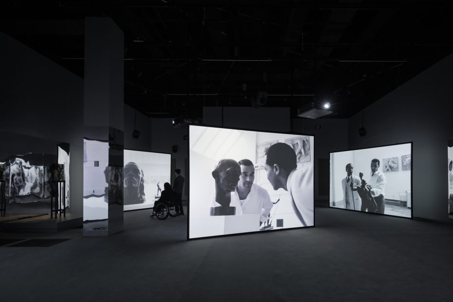A dark room with large illuminated screens displaying black-and-white images of people in conversation and a person in a wheelchair in the background.