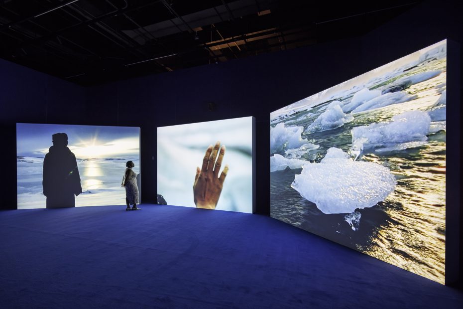 A person stands in a dark room looking at three illuminated screens displaying images of icebergs, a hand, and a silhouetted figure against a sunset over water.