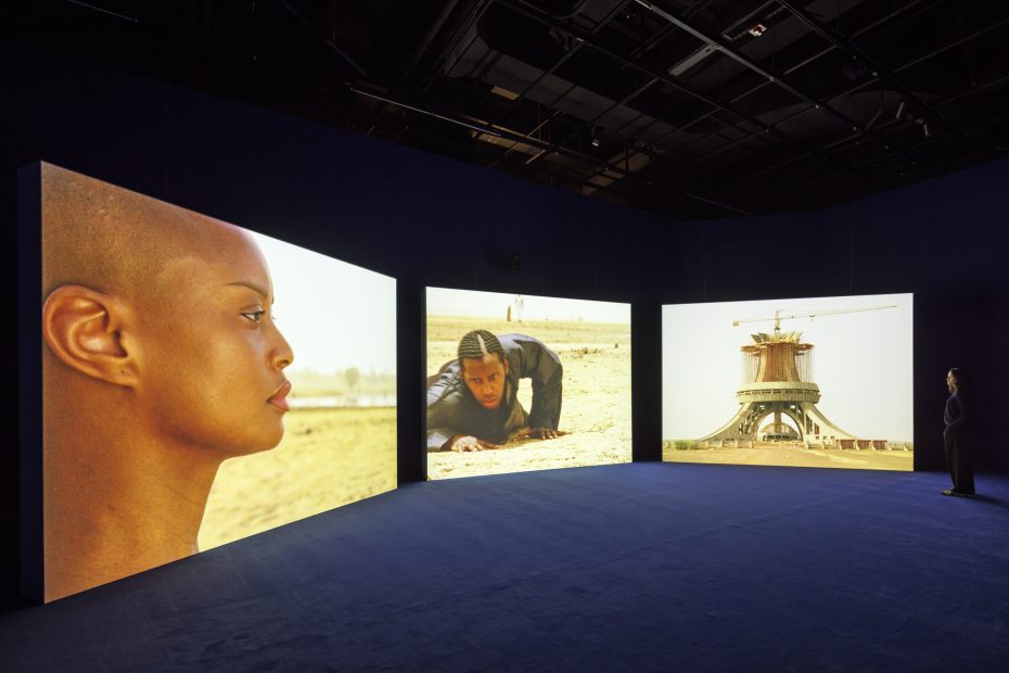 Three large screens display a close-up of a woman's face, a person crawling on sand, and a construction site, in a dark exhibition space with a visitor observing.