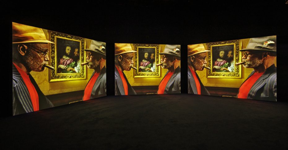 Three large projection screens display a man in a hat and glasses looking at a framed portrait painting in a dimly lit exhibition space.